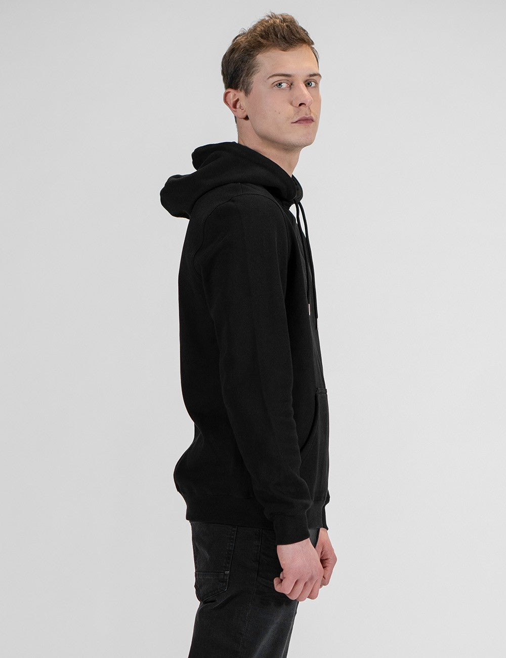 A side profile shot of a man wearing a plain black hoodie and dark jeans, standing against a clean, light grey background.