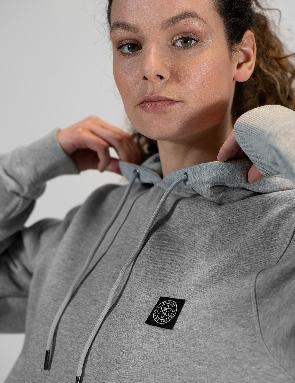 A close-up shot of a woman with curly hair tied back, wearing a heather grey pullover hoodie. She has her hands up near the collar, adjusting the hood. A small, square black logo patch featuring a circular white graphic is stitched on the left chest of the sweatshirt.