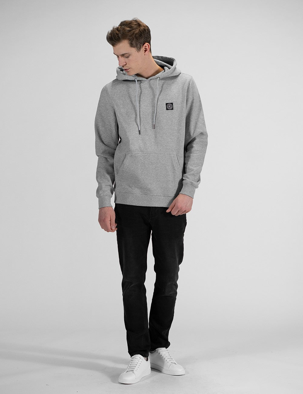 A man stands looking down and to his right, wearing a heather grey drawstring hoodie with a small rectangular logo patch on the left chest, black slim-fit jeans, and clean white sneakers.