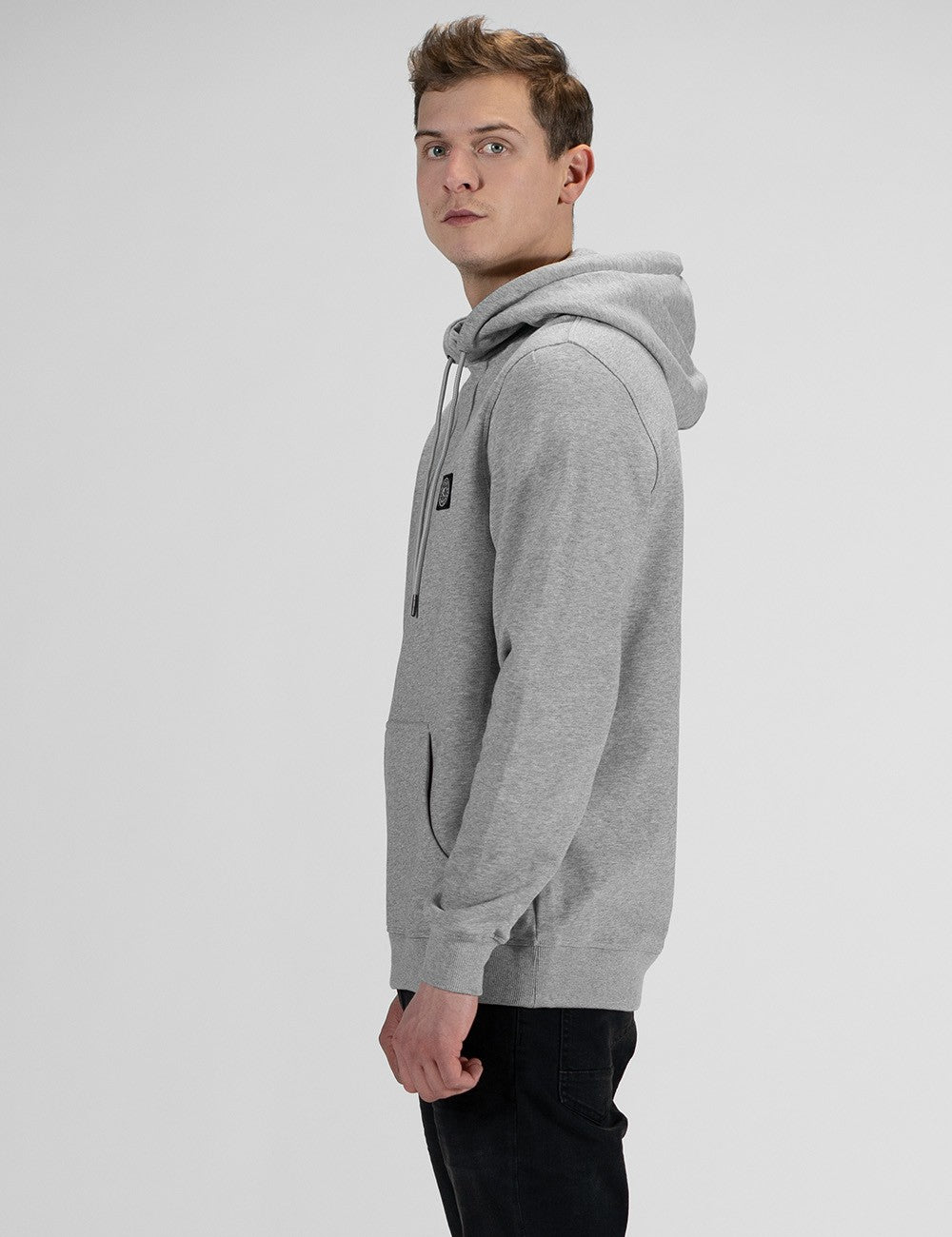 A young man with light brown hair stands in profile against a plain white background, looking back toward the camera over his shoulder. He is wearing a light grey hoodie with the hood up, drawstring cords visible, and dark pants.