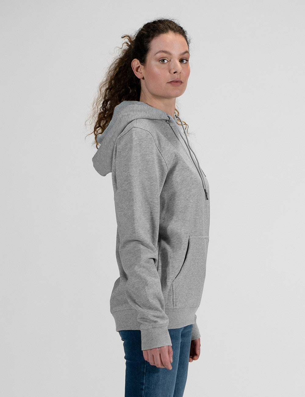 A side profile of a woman with long, curly brown hair wearing a grey hoodie and blue jeans against a plain white background.
