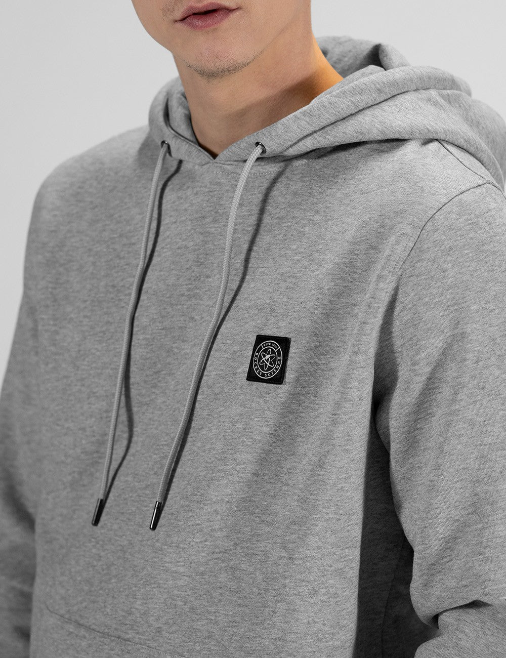 A close-up shot of a person wearing a light grey pullover hoodie. The focus is on the upper chest, showing two long grey drawstrings with metallic aglets hanging down from the hood. On the left side of the chest, there is a small, square black patch featuring a circular logo with a central symbol resembling an atom or a compass design. 