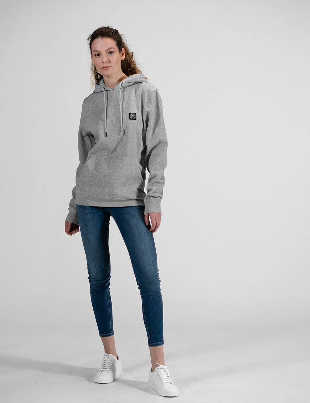 A woman with long brown hair stands facing forward, wearing a light grey hoodie with a small black square logo patch on the left chest, blue skinny jeans, and white sneakers.