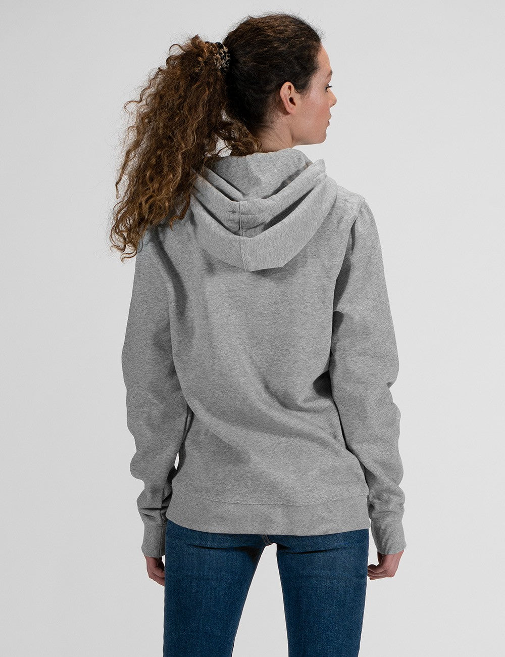A rear profile of a woman with long, curly brown hair wearing a grey hoodie and blue jeans .