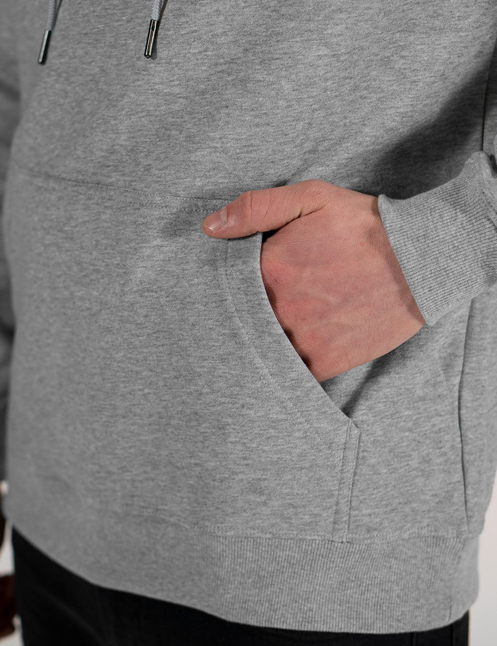 A close-up shot focuses on a person's hand resting inside the front kangaroo pocket of a light grey heathered hoodie. The hoodie features a ribbed hem at the bottom and visible black drawstrings near the top collar.
