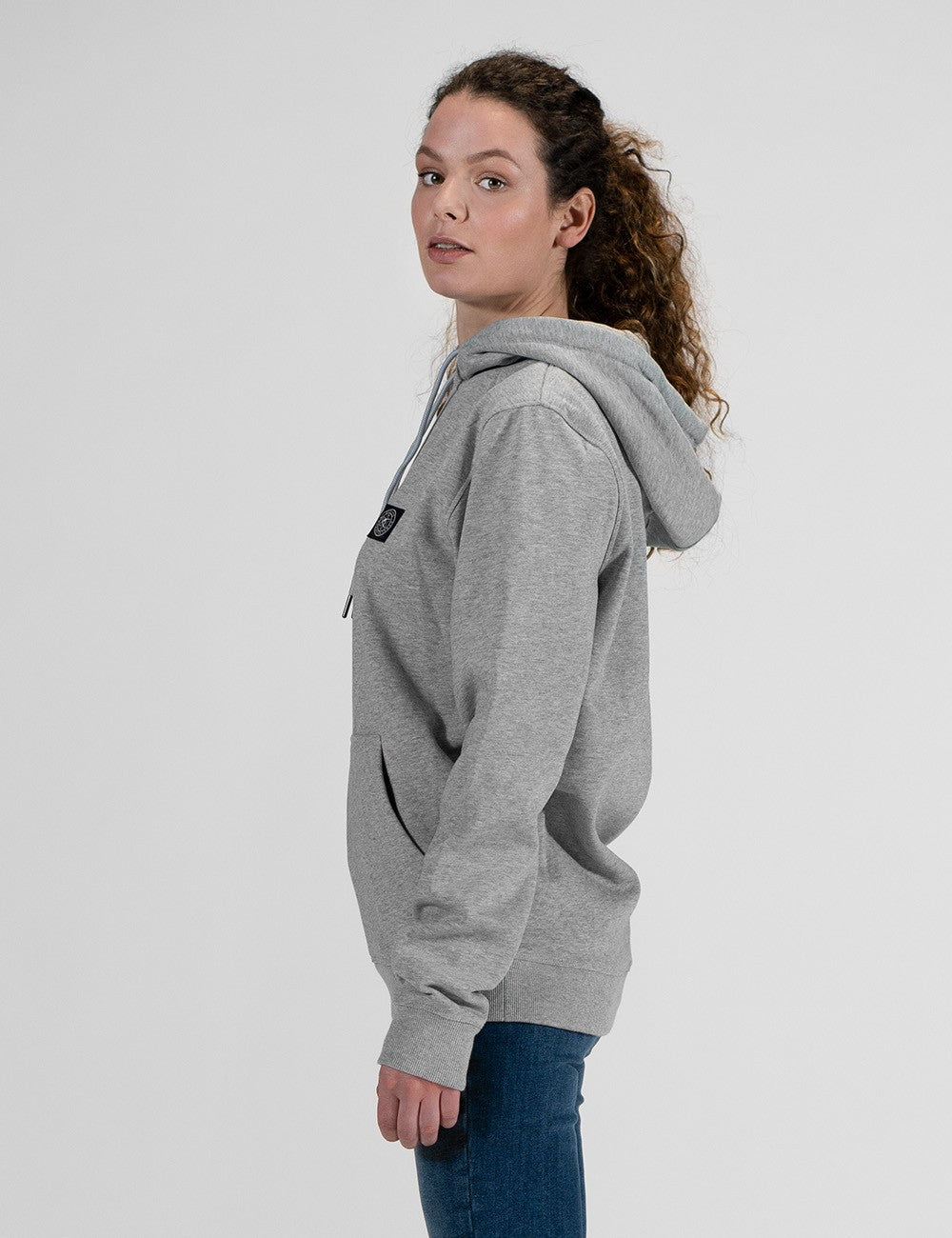 A side profile of a woman with long, curly brown hair wearing a grey hoodie and blue jeans .