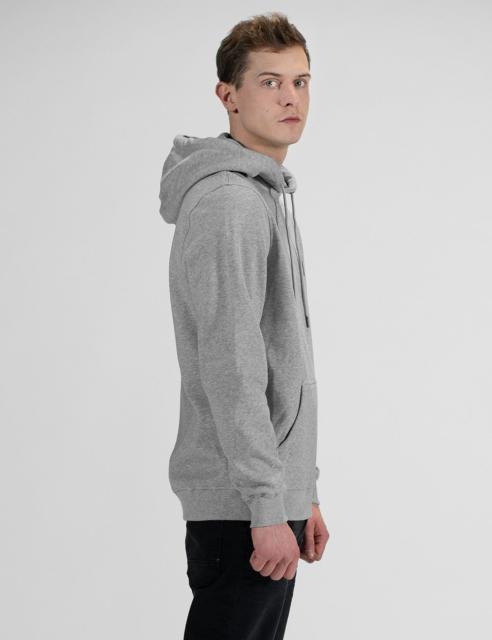 A young man with light brown hair stands in profile against a plain white background, looking back toward the camera over his shoulder. He is wearing a light grey hoodie with the hood up, drawstring cords visible, and dark pants.
