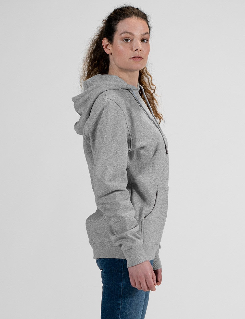 A studio shot of a person wearing a premium light heather grey pullover hoodie. The hoodie features a relaxed, medium-fit design with a matching grey drawstring hood, a classic front kangaroo pocket.