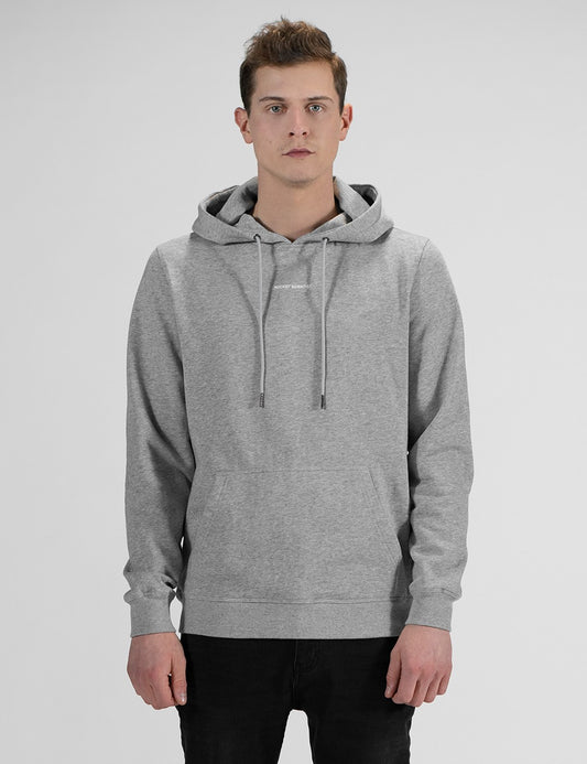 A man standing in a front-profile view against a plain, light grey background, wearing a heather grey pullover hoodie , with a small print in he chest and black pants.