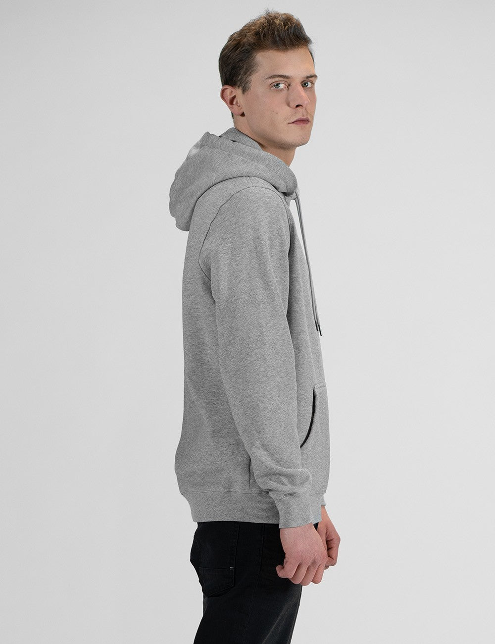 A man standing in a side-profile view against a plain, light grey background, wearing a heather grey pullover hoodie and black pants.