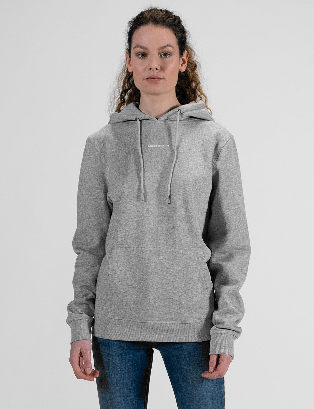 A front-facing, medium shot shows a woman with curly hair tied back wearing a grey pullover hoodie with drawstrings and a central pouch pocket, paired with blue jeans, set against a plain light grey background.