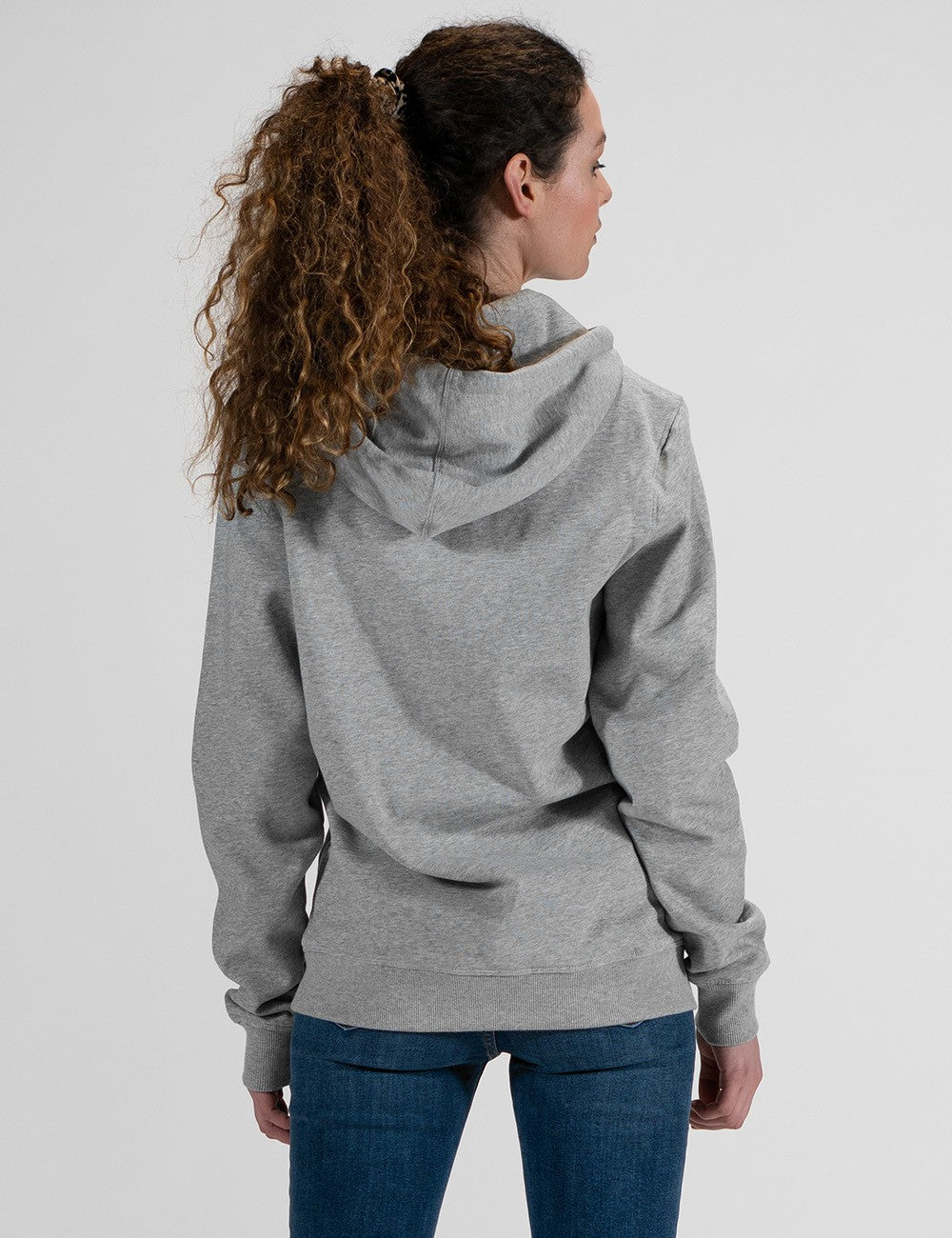 A woman with long, curly brown hair tied back in a high ponytail is viewed from behind. She is wearing a light grey pullover hoodie with the hood down, and slim-fit blue jeans.