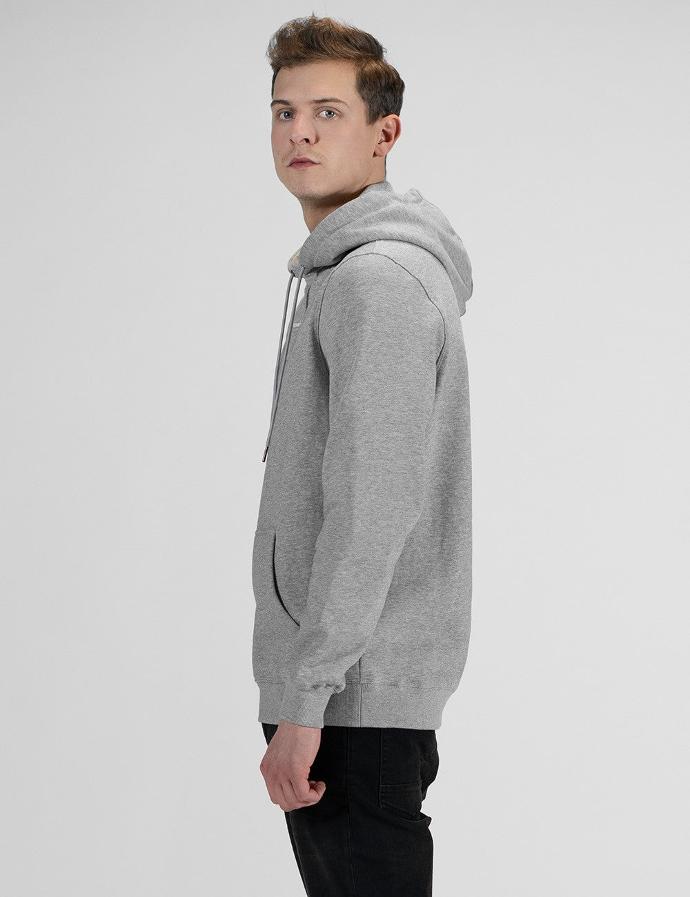 A man standing in a side-profile view against a plain, light grey background, wearing a heather grey pullover hoodie and black pants.