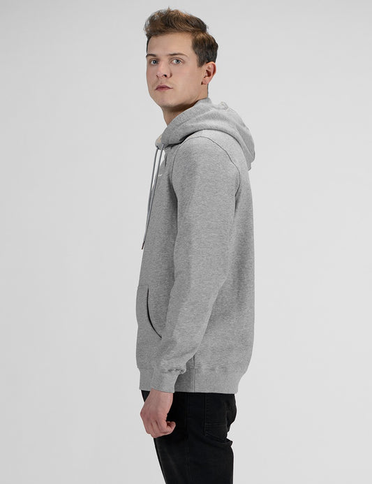 A man standing in a side-profile view against a plain, light grey background, wearing a heather grey pullover hoodie and black pants.