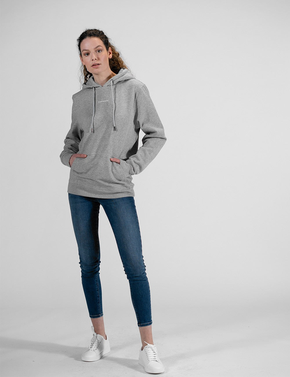 A studio shot of a person wearing a premium light heather grey pullover hoodie. The hoodie features a relaxed, medium-fit design with a matching grey drawstring hood, a classic front kangaroo pocket, and small, minimalist white text branding printed subtly across the center chest.