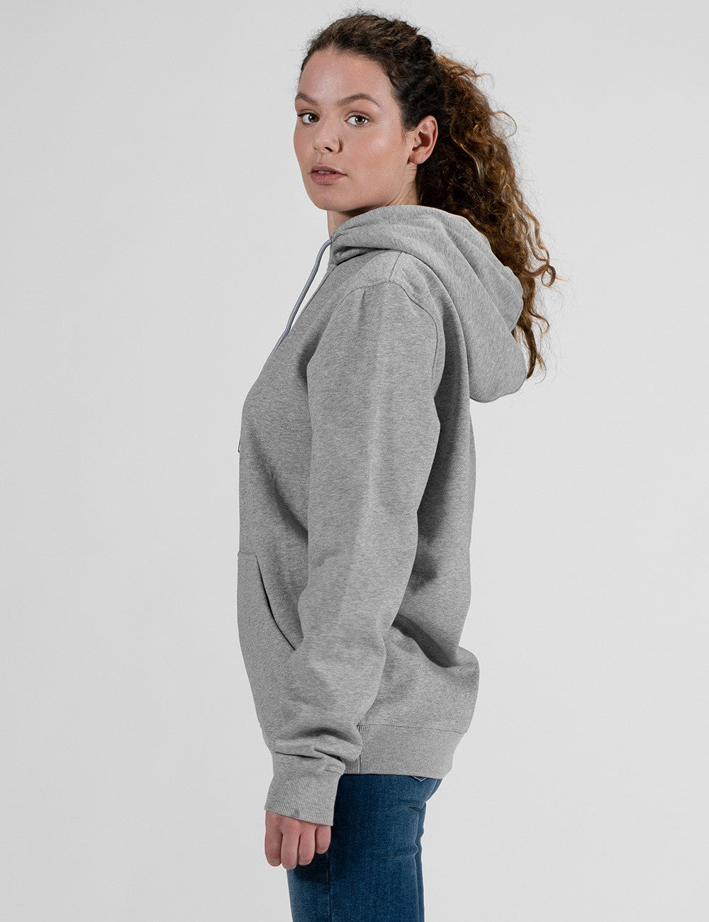 A woman with long, curly brown hair tied back in a high ponytail is viewed from the side. She is wearing a light grey pullover hoodie with the hood down, and slim-fit blue jeans.