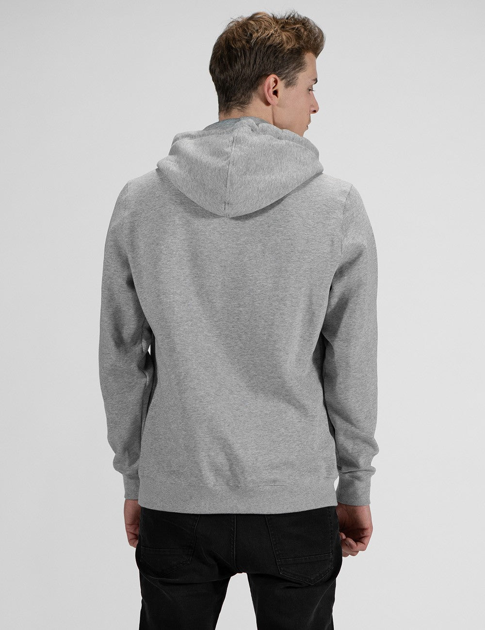 A man standing in a rear-profile view against a plain, light grey background, wearing a heather grey pullover hoodie and black pants.