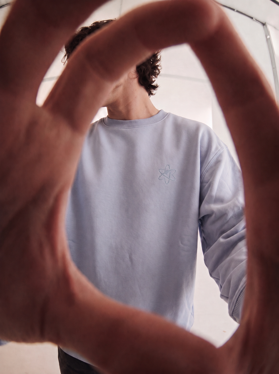A person wearing a light periwinkle blue crewneck sweatshirt with a small embroidered atom symbol on the left chest, framed by a large hand in the foreground.