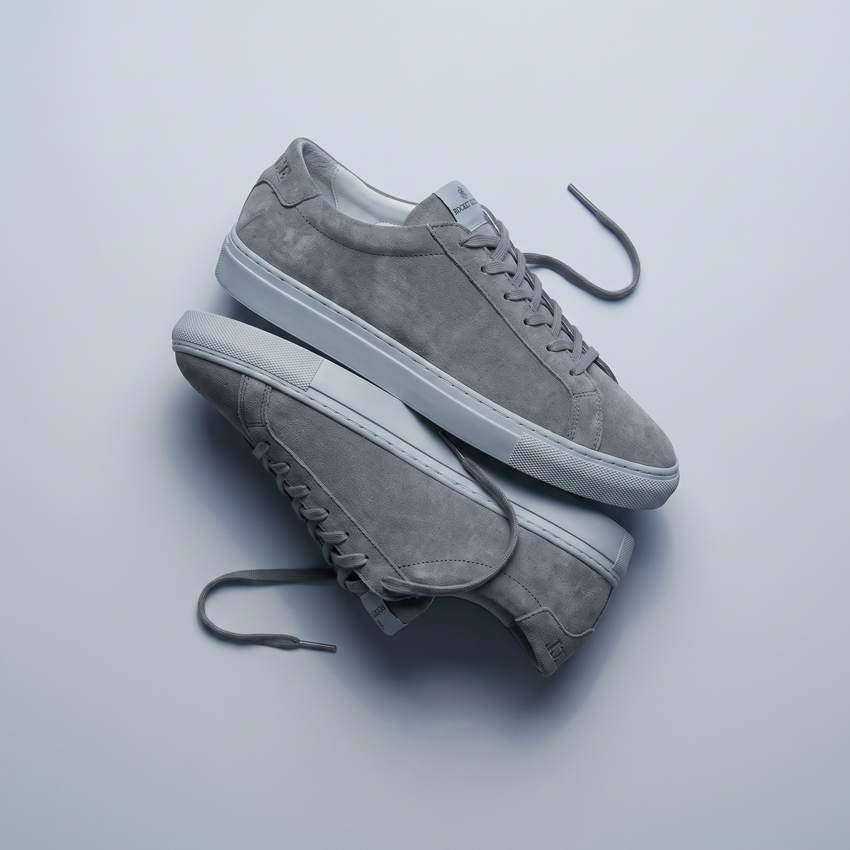 A pair of low-top gray suede sneakers with white rubber soles, arranged diagonally on a light gray background.