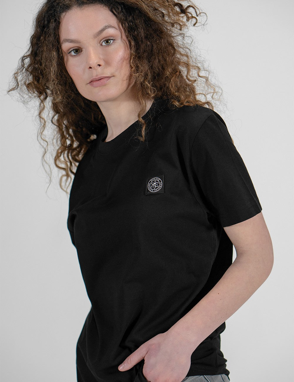A woman with long, curly brown hair wearing a plain black crewneck t-shirt with a small circular logo patch on the left chest.