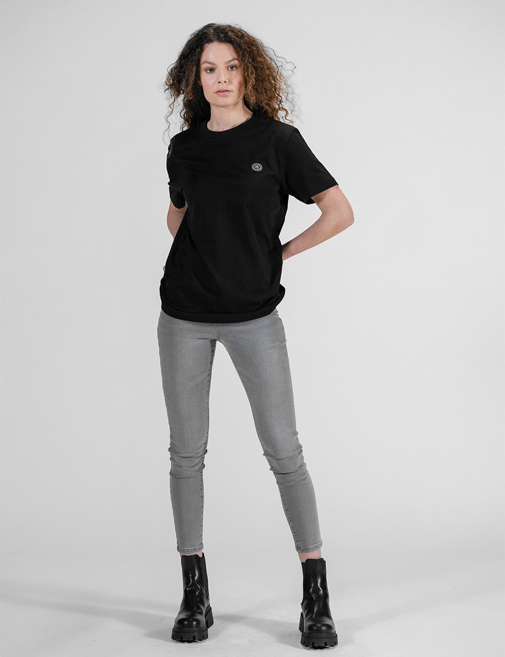 A woman with curly hair poses in a black t-shirt, slim grey pants, and chunky black boots against a plain grey background.