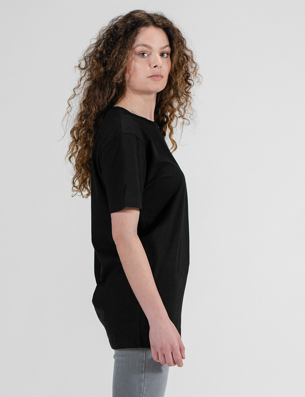 A woman with long, curly brown hair stands facing to the right, showing the side profile of a plain, solid black short-sleeve t-shirt.