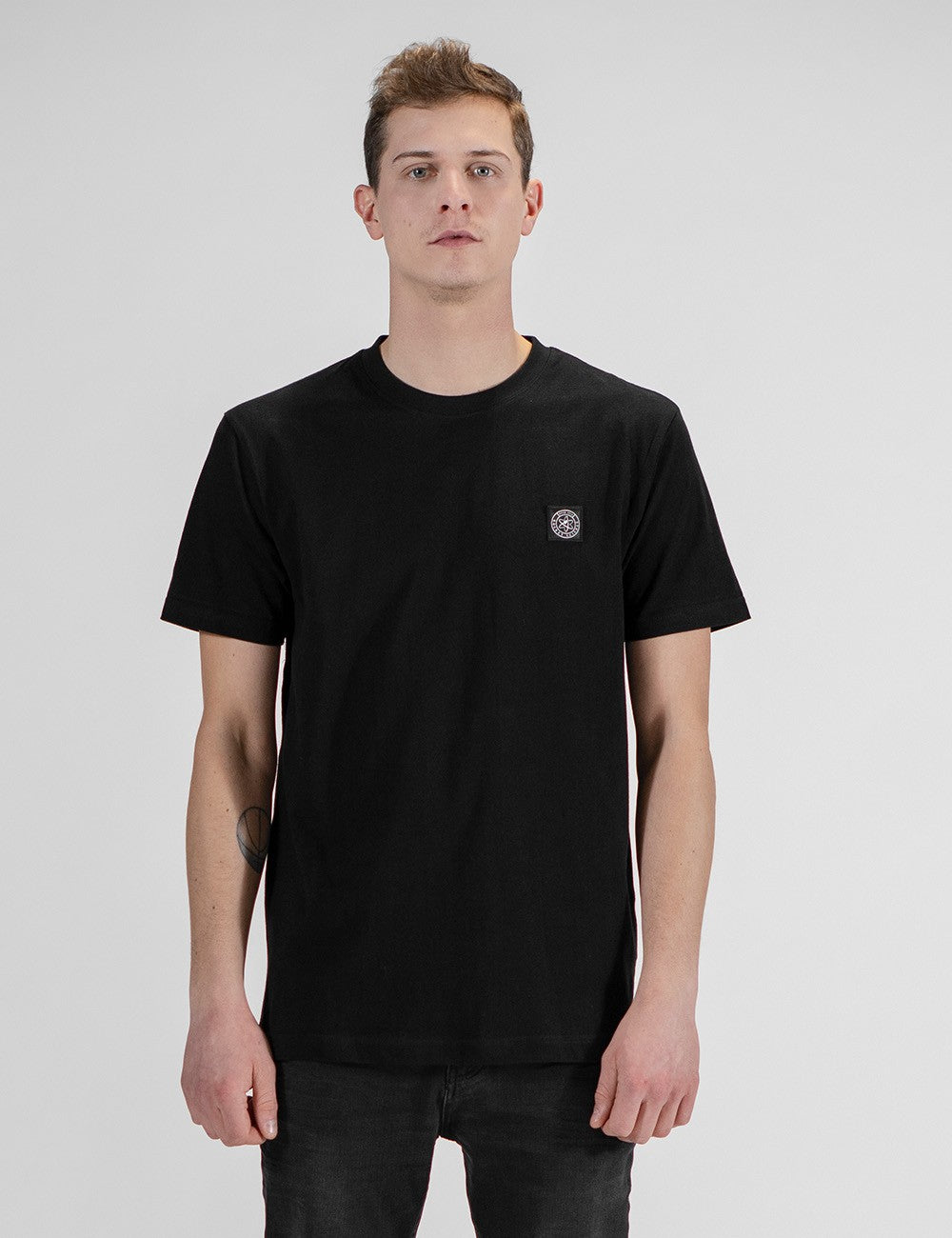 This image shows a front pose of young man wearing a black cotton crew-neck t-shirt , with a patch logo on the left side.
