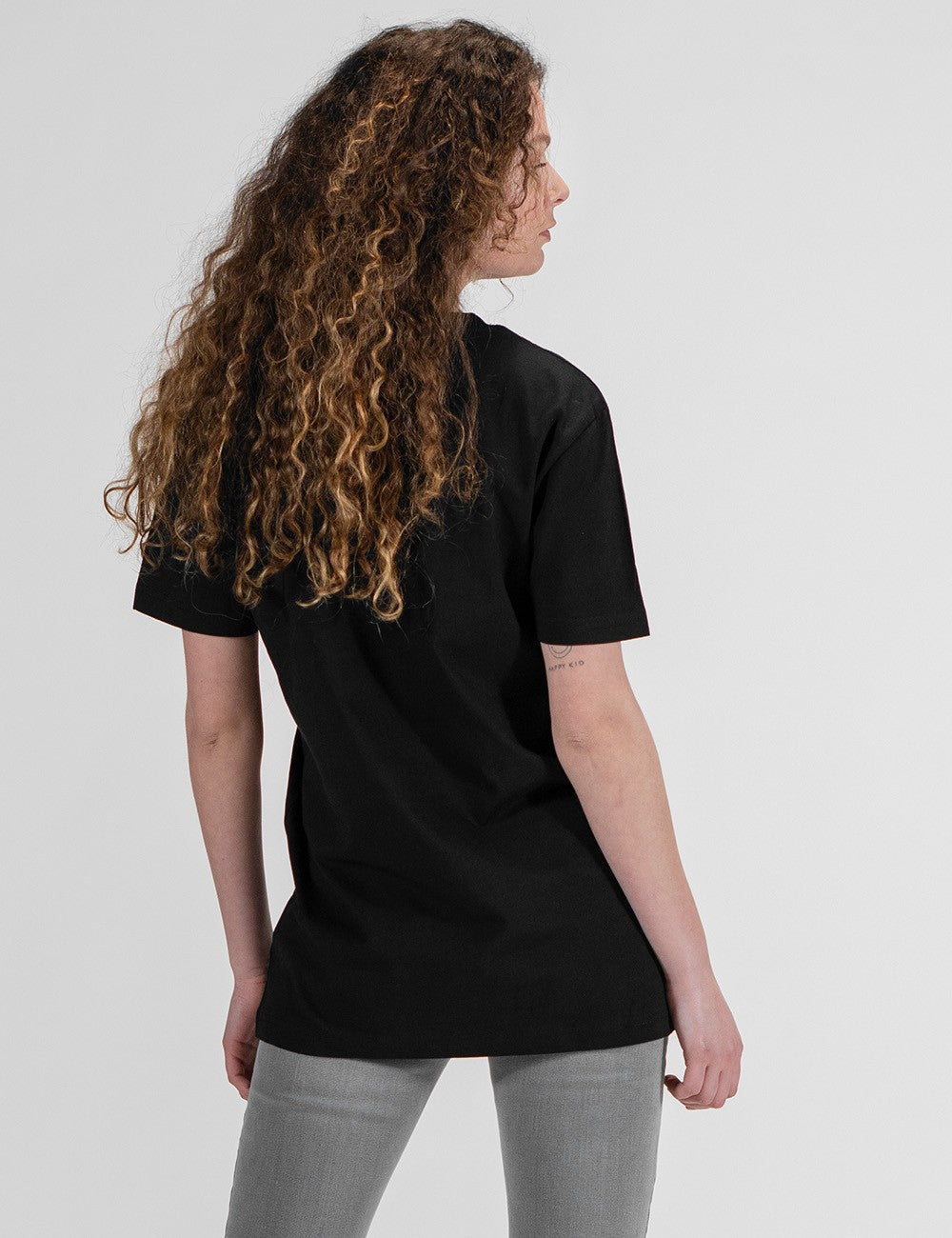 A woman with long, curly blonde and brown hair is seen from behind, looking to her right. She is wearing a plain black short-sleeved t-shirt and grey skinny jeans.