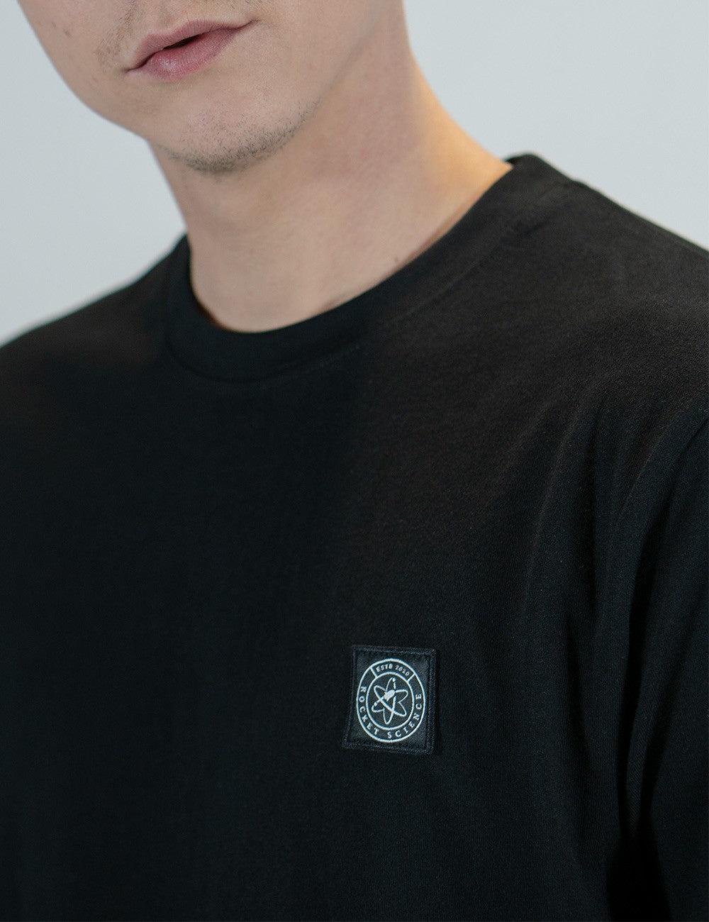 A close-up shot of a person wearing a plain black crew-neck t-shirt. The shirt features a small, square black patch on the left chest area, which displays a white circular logo.