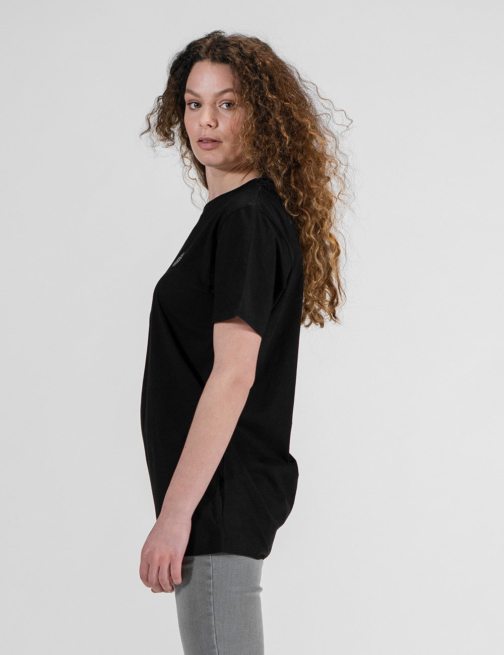 A woman with long, curly brown hair stands facing to the right, showing the side profile of a plain, solid black short-sleeve t-shirt.
