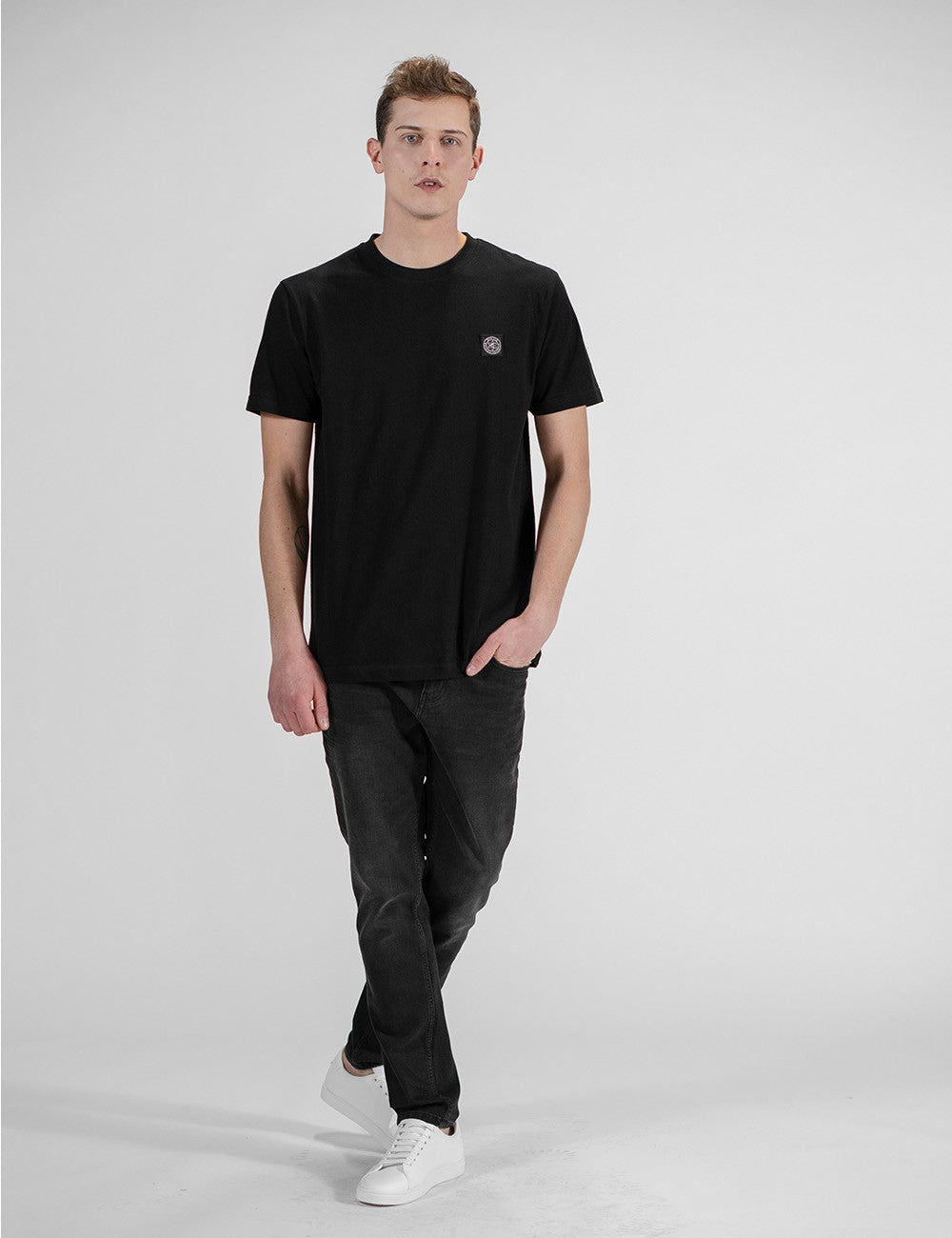 This image shows a young man wearing a black cotton crew-neck t-shirt with a small chest logo patch, paired with dark grey jeans and white sneakers.