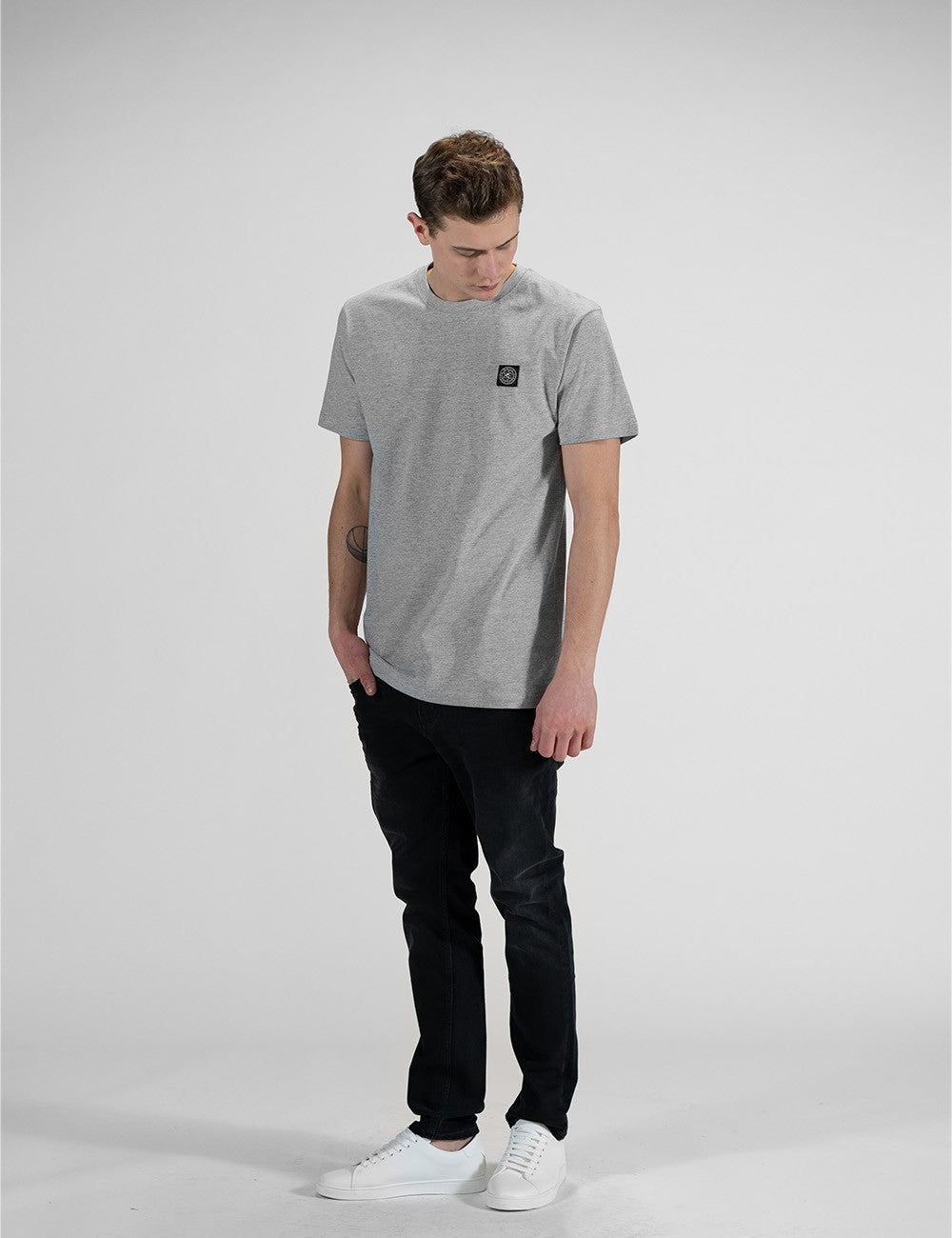 A young man with light brown hair, wearing a light grey Rocket Science short-sleeve t-shirt with a small square compass logo patch on the left chest.