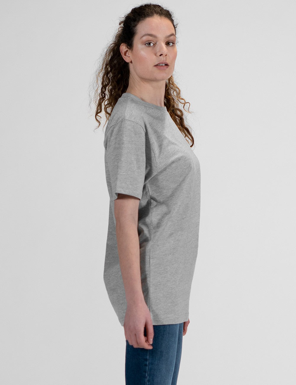This image shows a side pose of young woman wearing a basic heather grey crew neck t-shirt and blue jeans.