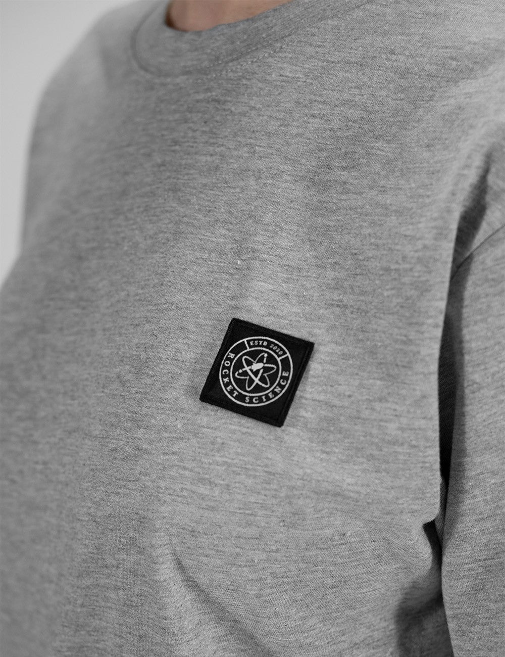 The image shows a grey cotton crewneck top featuring a round neckline and a square badge Rocket Science logo patch on the chest.