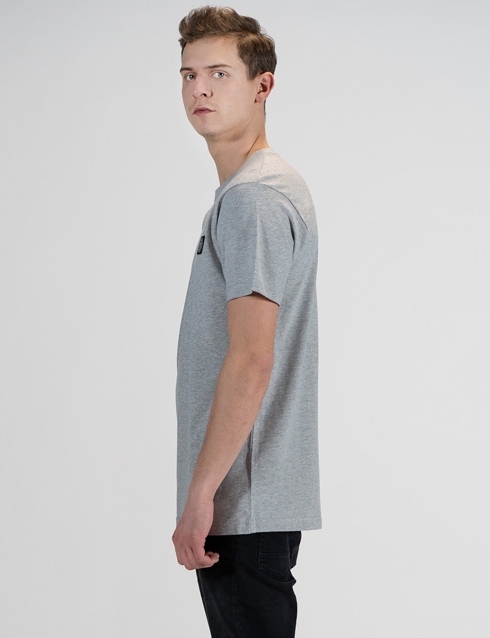 This image shows the side pose of a young man with light brown hair, wearing a light grey Rocket Science short-sleeve t-shirt with a small square compass logo patch on the left chest.
