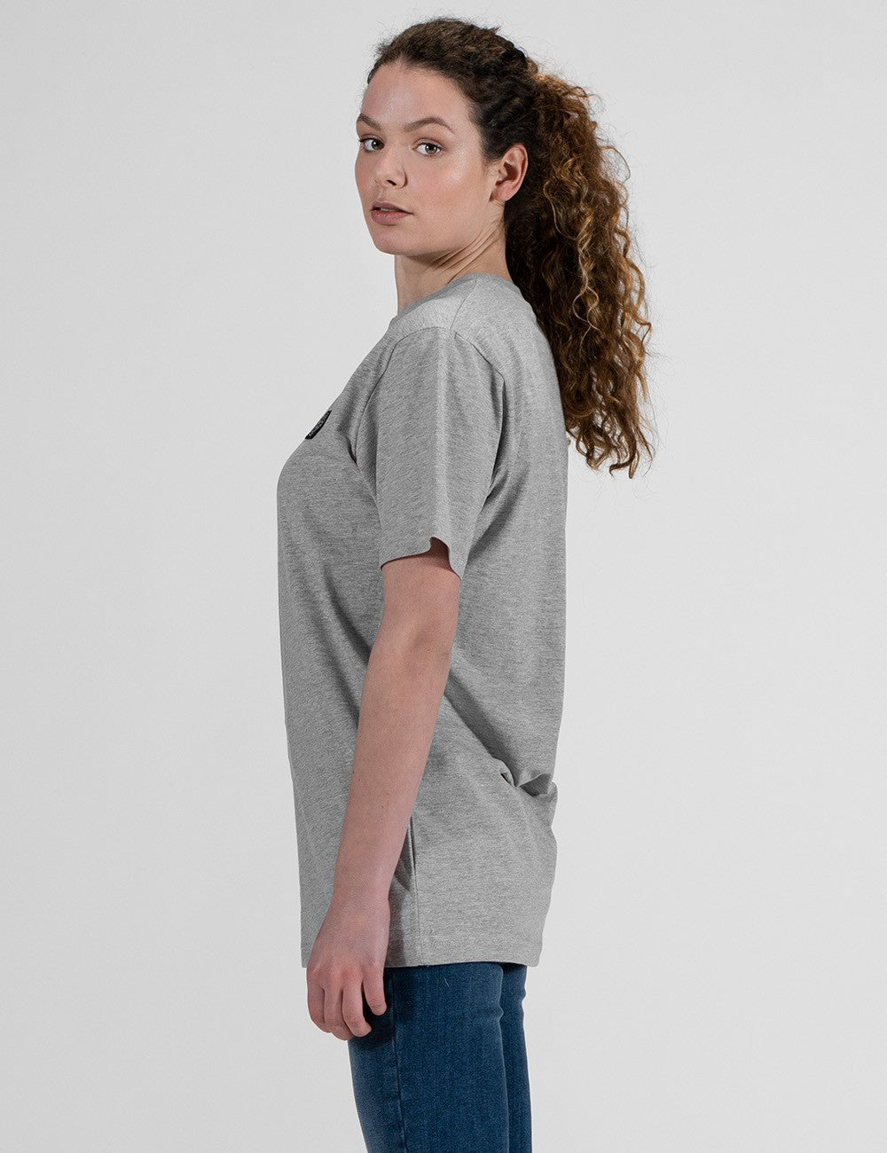 This image shows a side pose of young woman wearing a basic heather grey crew neck t-shirt and blue jeans.