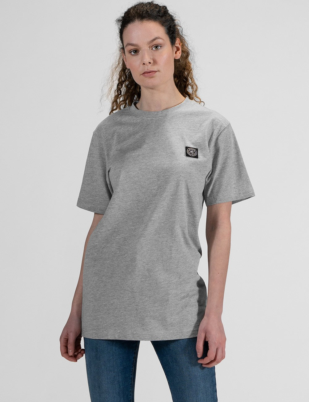 This image shows a front pose of young woman wearing a basic heather grey crew neck t-shirt with a square patch logo on the left side and blue jeans.