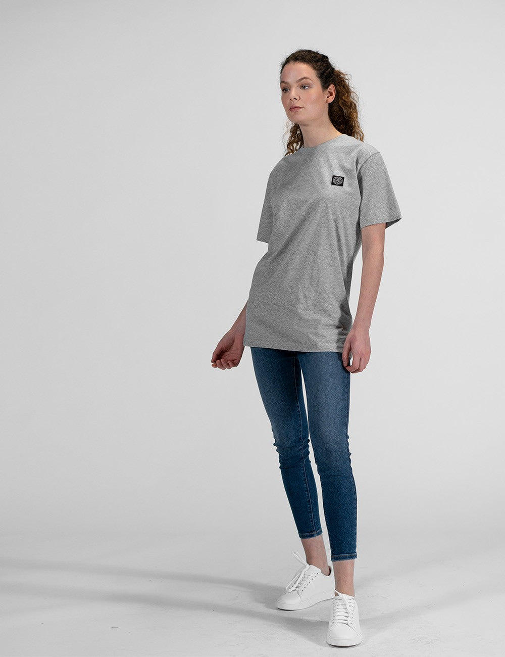 A woman with long, curly brown hair stands against a plain light background, looking slightly away from the camera. She is wearing an oversized heather grey short-sleeved t-shirt with a small square black graphic patch on the left side of the chest. She has paired the shirt with dark blue skinny ankle-length jeans and plain white leather low-top sneakers.