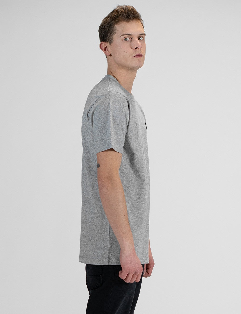 This image shows the side pose of a young man with light brown hair, wearing a light grey Rocket Science short-sleeve t-shirt with a small square compass logo patch on the left chest.
