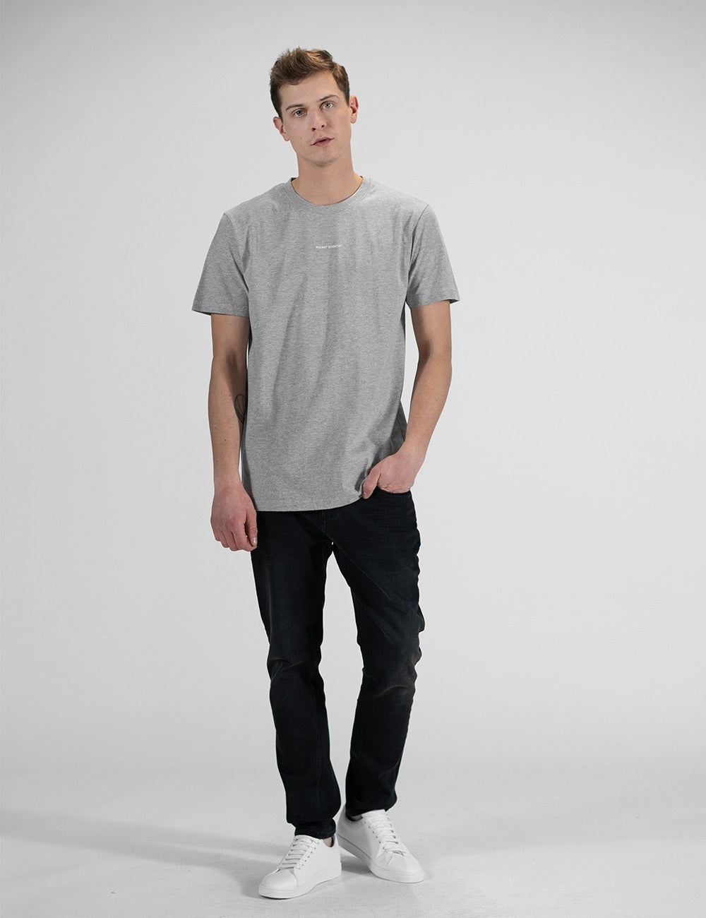 This image shows a man wearing a casual grey crew-neck t-shirt, dark denim jeans, with a chest print, and white sneakers.