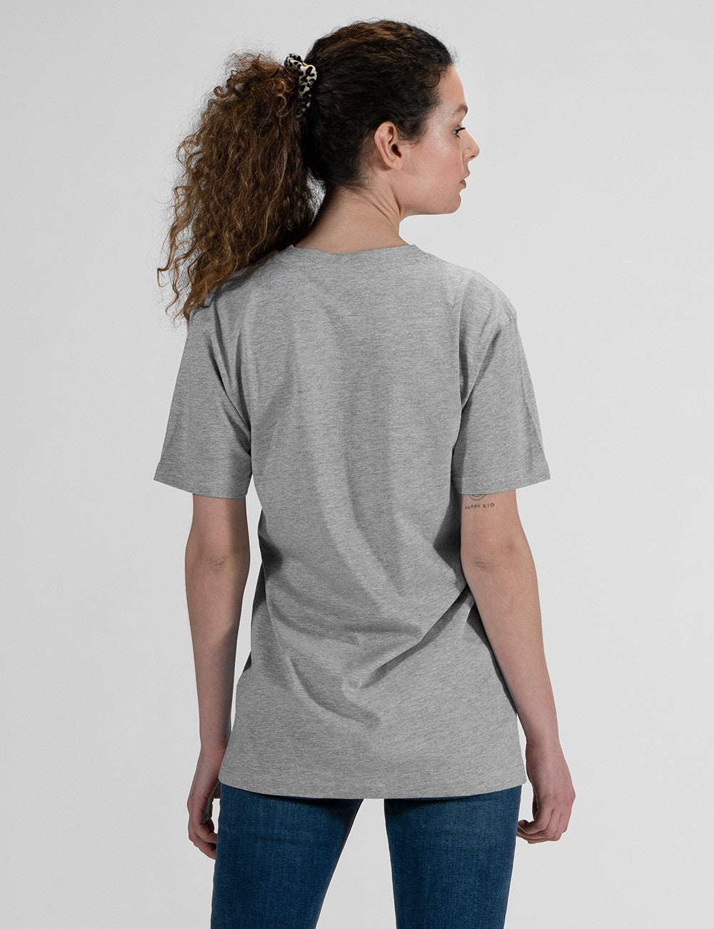 This image shows a rear pose of a young woman wearing a grey crew-neck t-shirt, dark blue jeans,