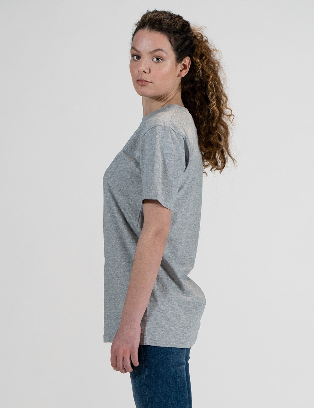 This image shows a side pose of a young woman wearing a grey crew-neck t-shirt, and dark blue skinny jeans.