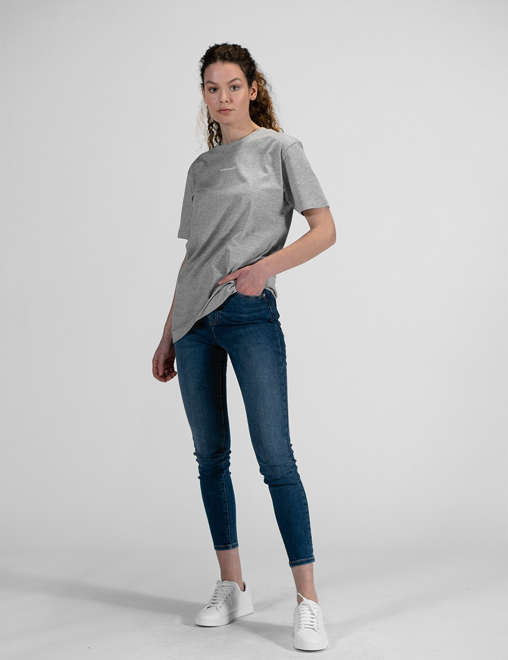 This image shows a young woman wearing a grey crew-neck t-shirt, dark blue skinny jeans, and white sneakers.