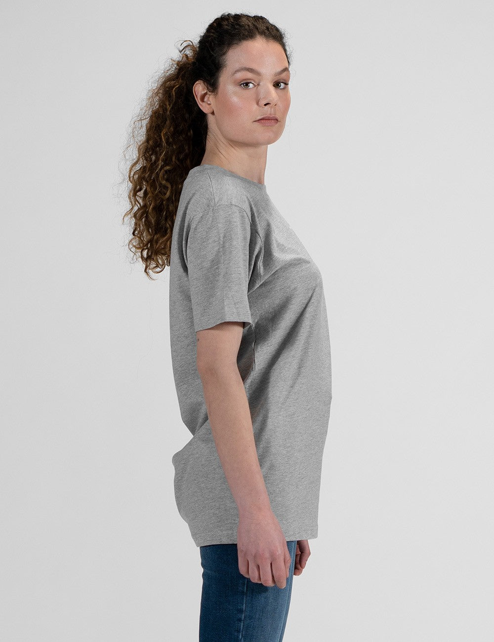 This image shows a side pose of a young woman wearing a grey crew-neck t-shirt, dark blue skinny jeans.