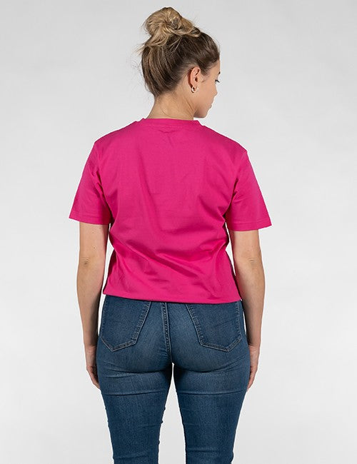 A woman viewed from behind wearing a bright pink short-sleeve crop top and blue jeans against a light gray studio background.