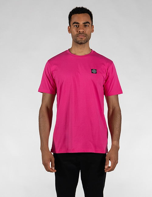A man wearing a bright pink crewneck t-shirt with a small black and white Rocket Science logo on the left chest, paired with black pants, standing against a light gray studio background.