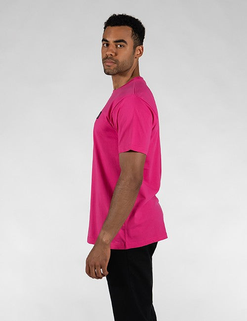 A man in profile wearing a bright pink short-sleeve crewneck t-shirt and black pants, standing against a light gray studio background.