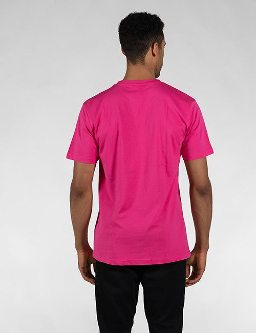 A man viewed from behind wearing a bright pink short-sleeve t-shirt and black pants, standing against a light gray studio background.