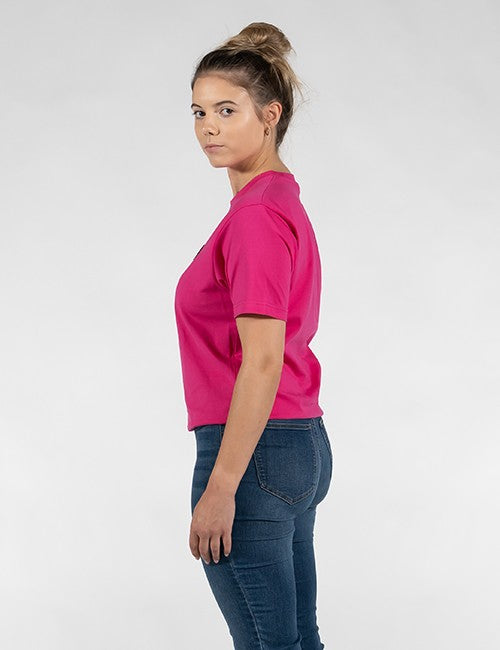 A woman shown from the side profile wearing a bright pink short-sleeve crewneck t-shirt and dark blue jeans, standing against a light gray studio background.