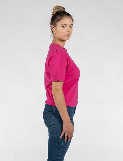 A woman shown in side profile wearing a bright pink short-sleeve crewneck t-shirt and dark blue jeans, standing against a light gray studio background.